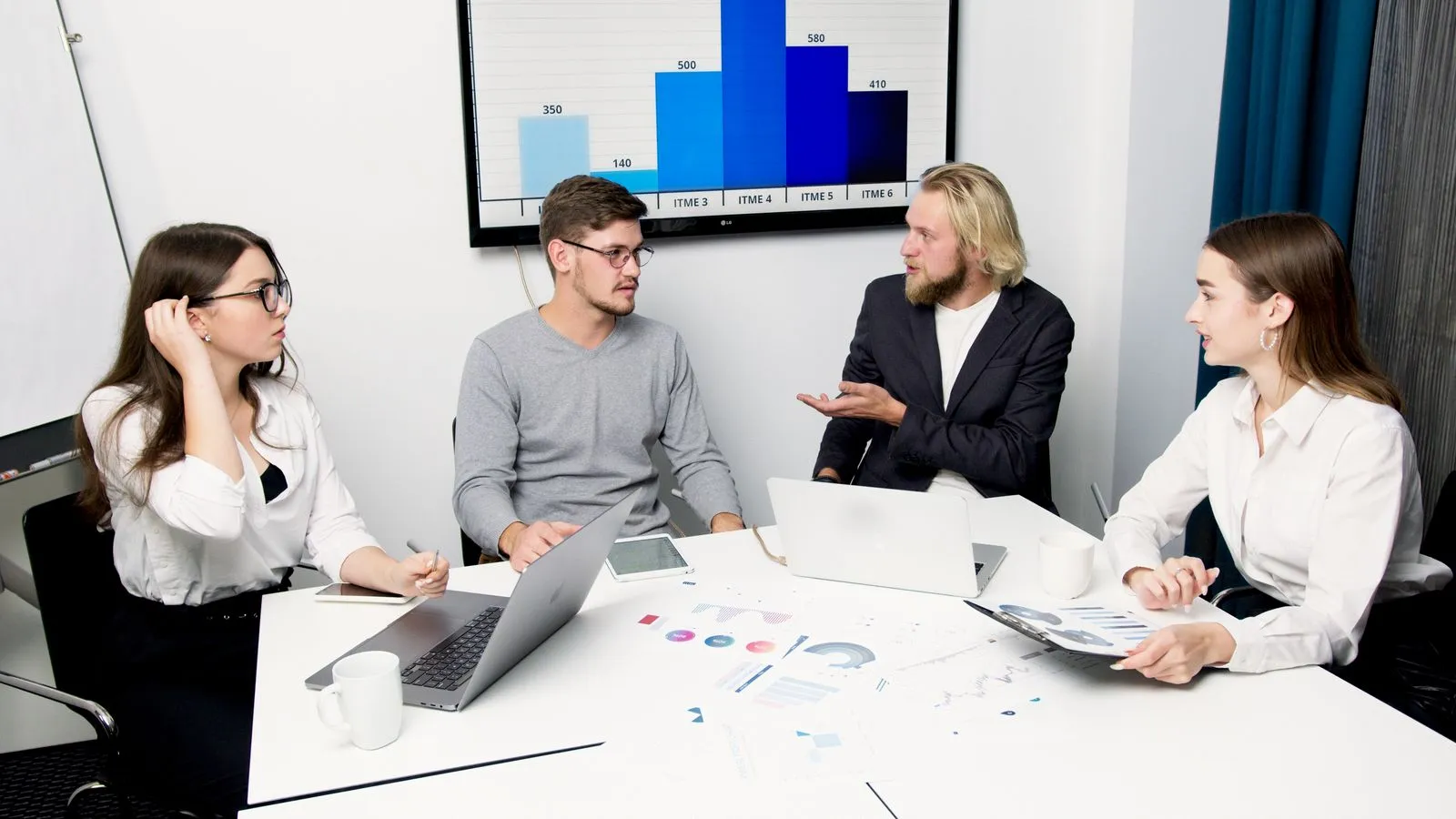 A team of four discussing results around a table with laptops and a bar chart on the wall screen