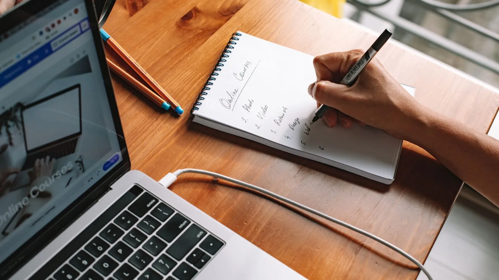 A person takes notes in a notebook while following an online course on a laptop