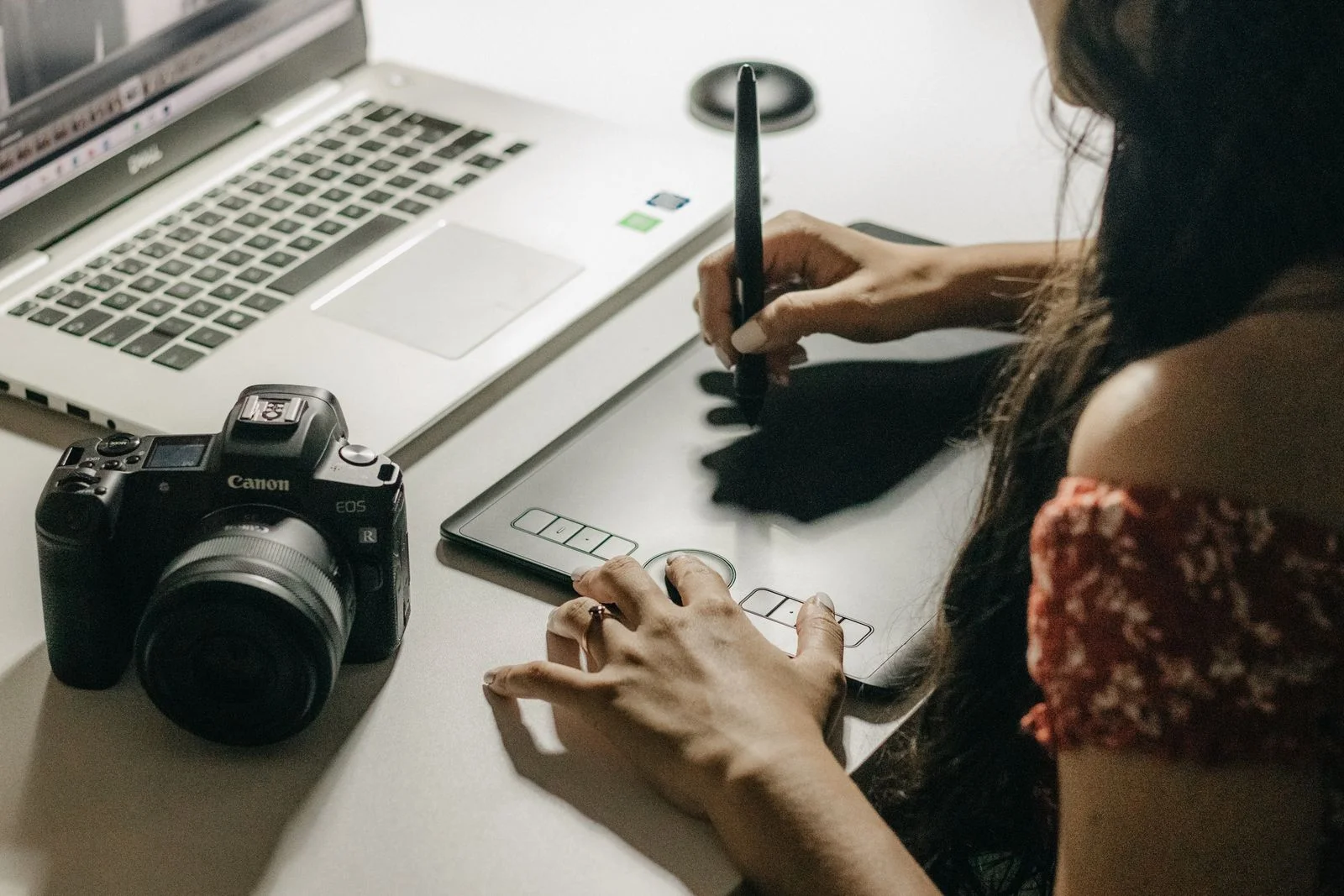 A designer works with a stylus on a graphics tablet next to a laptop and a camera