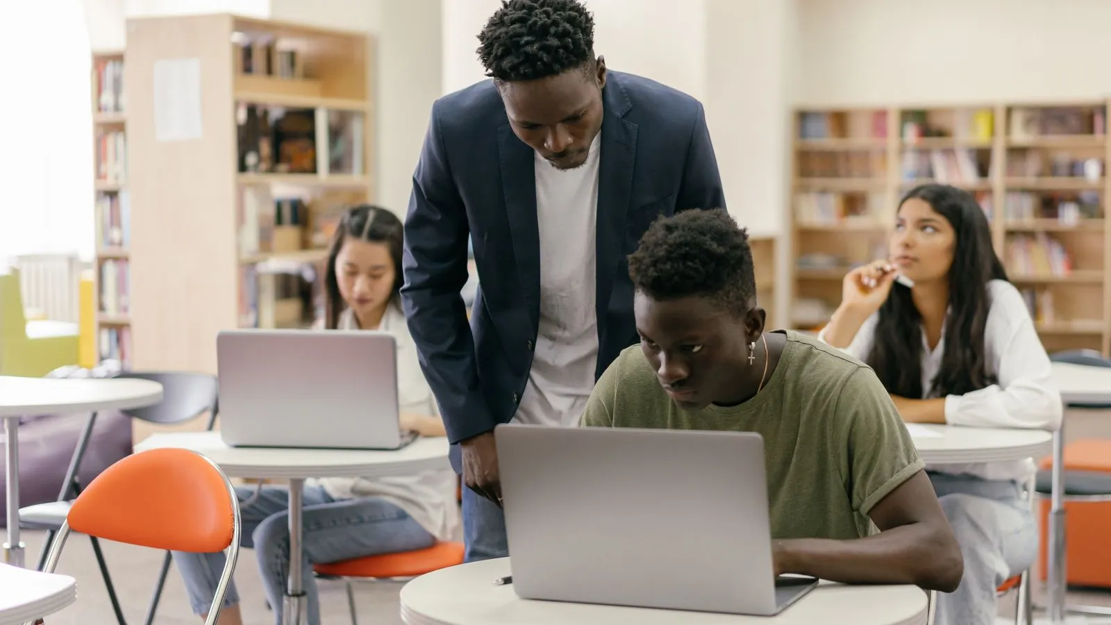 A teacher leans over a student working on a laptop in a classroom