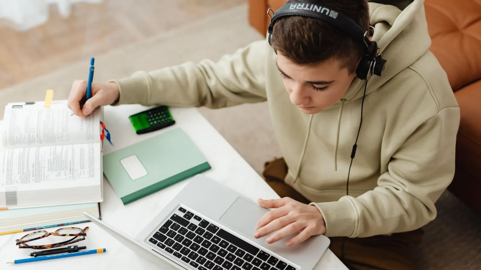 A teenager does homework at a desk with a laptop, a textbook and a notebook