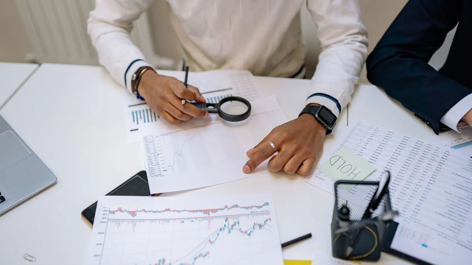 Two people review financial charts and documents on a desk with a magnifying glass