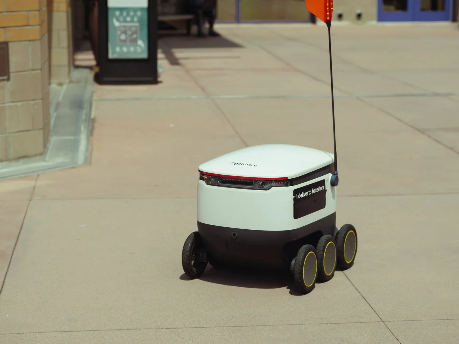 A six-wheeled sidewalk delivery robot with a safety flag crossing a paved university walkway