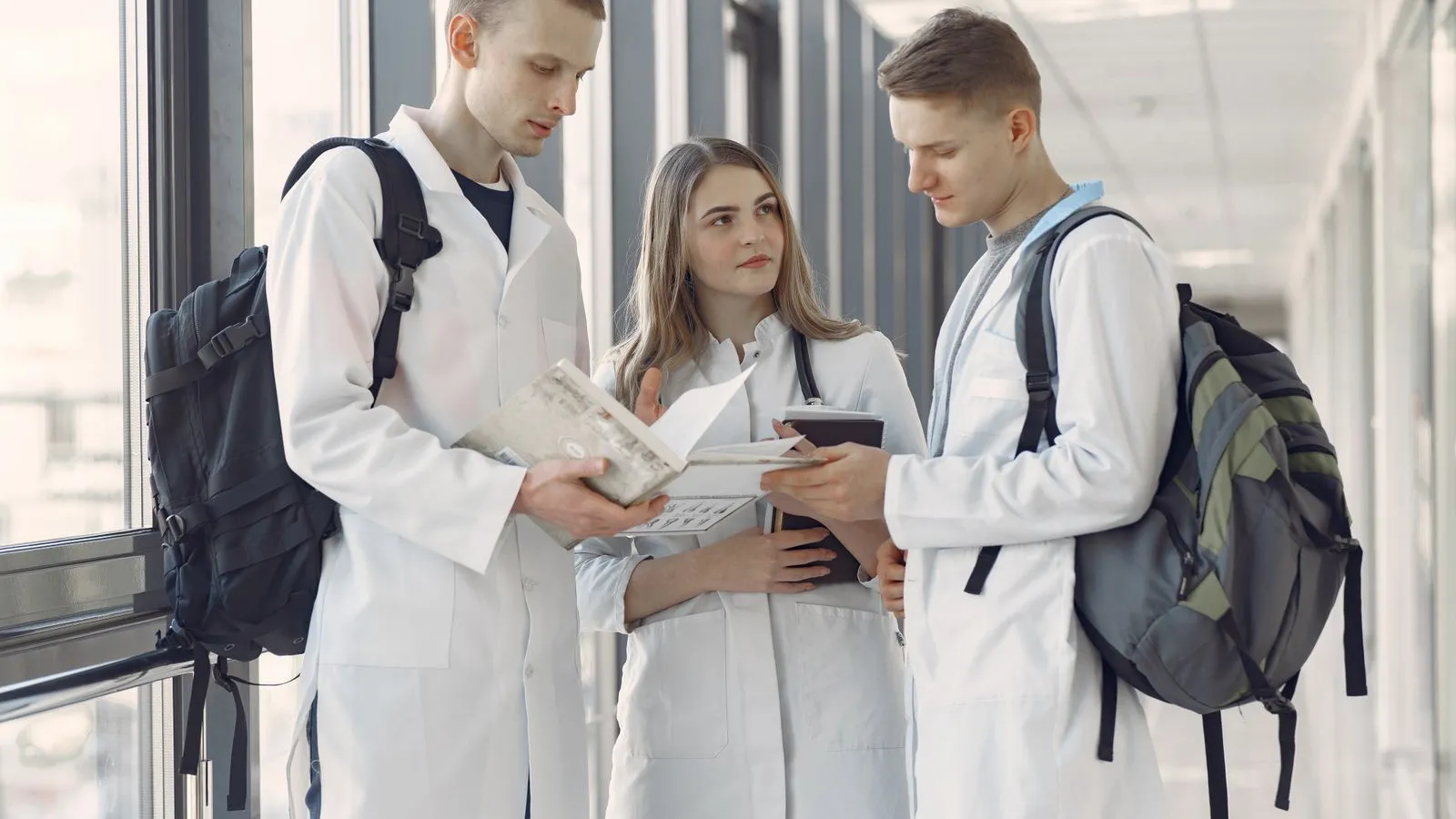 Three medical students in white coats reviewing notes in a hospital corridor